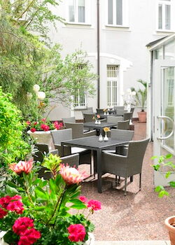 Charmanter Innenhof mit modernen Rattanmöbeln, gedeckten Tischen und umgeben von blühenden Pflanzen und viel Grün. Zugang durch eine helle Glastür vom Wintergarten aus.