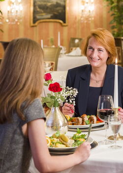 Zwei Frauen genießen ein elegantes Abendessen bei Kerzenschein in einem klassisch dekorierten Restaurant mit roten Wänden, goldenen Stühlen und Kronleuchtern. Auf dem Tisch stehen Wein, Wasser, ein Blumengesteck und festlich angerichtete Speisen.