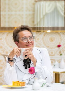 Eine Dame mit Brille sitzt in einem hellen Café mit einer Tasse in der Hand und vor ihr ein leckerer Kuchen auf dem Teller.