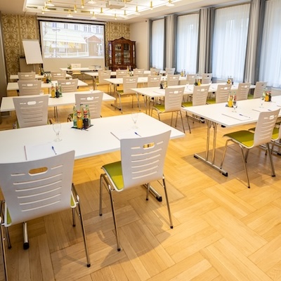 Ein heller Seminarraum mit mehreren Tischen, Stühlen und einer großen Leinwand.